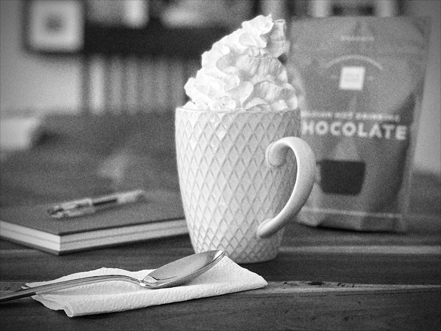 White mug with whipped cream on top of Cocoa Metro Organic Hot Chocolate on a table next to a notebook and a blurred package of Cocoa Metro's organic Belgian hot chocolate mix.