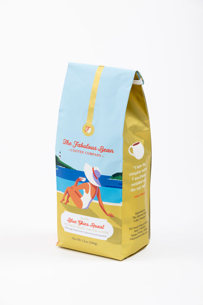 Specialty-grade medium light ground coffee blend from Central and South America in a colorful 12 ounce bag with the front panel depicting a sophisticated lady wearing a modest one piece bathing suit and floppy hat enjoying a cup of coffee while seated on a beautiful beach against a background of  light blue sky, green covered hills, an azure sea and golden sand. 