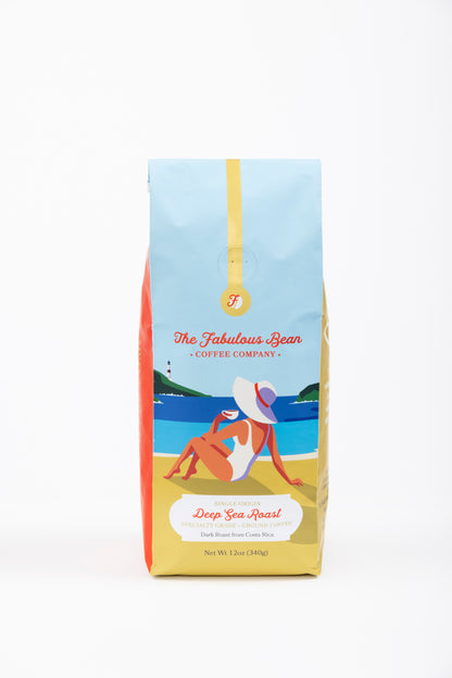 Specialty-grade dark roast ground coffee from Costa Rica in a colorful 12 ounce bag with the front panel depicting a sophisticated lady in a modest one piece bathing suit and floppy hat enjoying a cup of coffee on a beautiful beach against a background of  light blue sky, green covered hills, an azure sea and golden sand. 