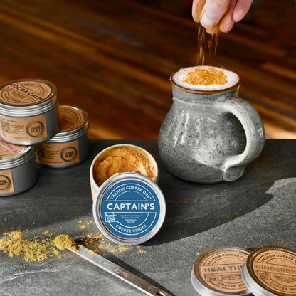 Captain's flavor coffee dust being added by hand to a large mug of hot chocolate. A full-size tin of Captain's coffee dust and several other flavors are depicted nearby on a slate table top.