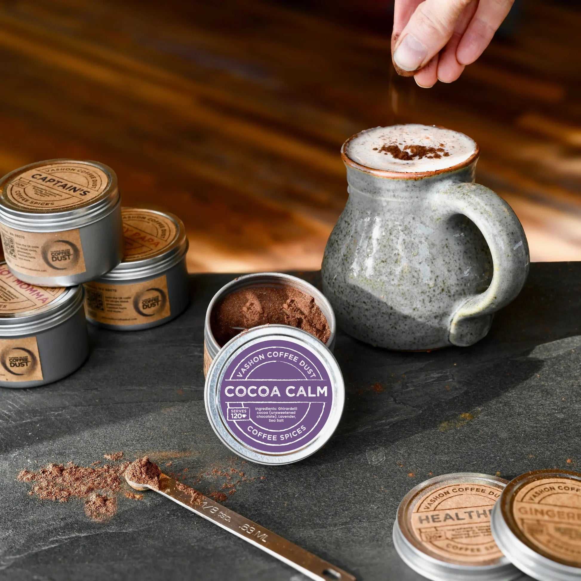 Vashon Island Coffee Dust Cocoa Calm flavor coffee dust full-size tin depicted with tins of other flavors and a hand sprinkling coffee dust onto a large mug of coffee on a wooden surface.