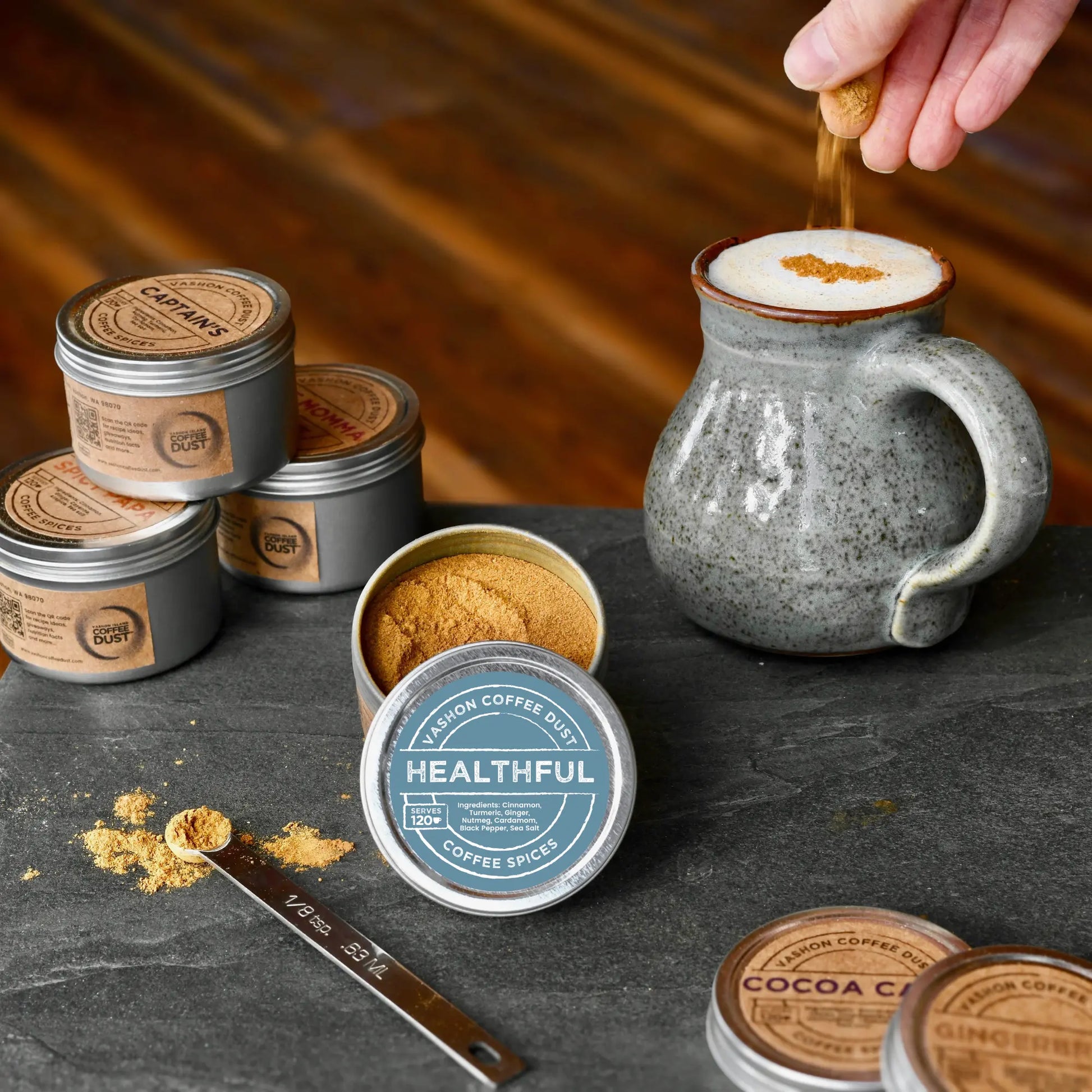 Healthful flavor coffee dust being added by hand to a large mug of hot chocolate. A full-size tin of Healthful coffee dust and several other flavors are depicted on a slate table top.
