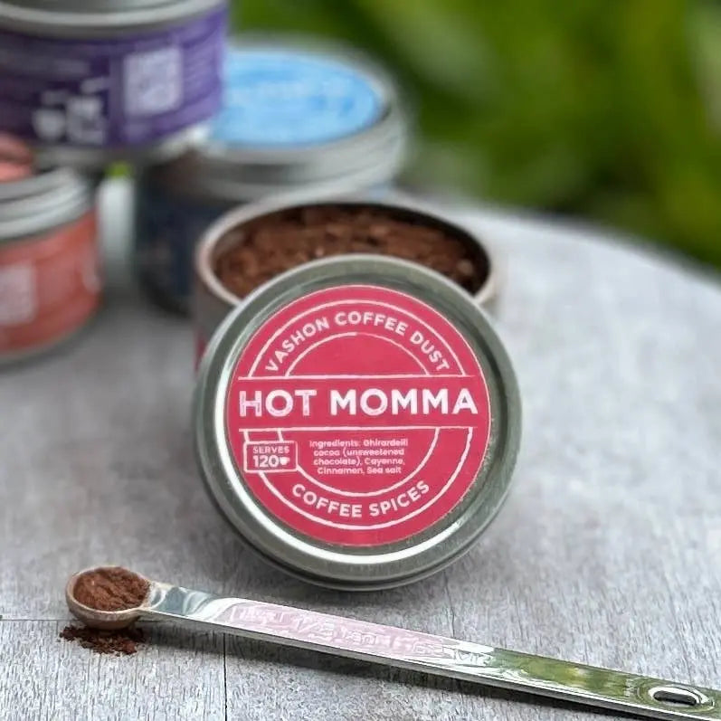 Tin of Vashon Island Coffee Dust flavor 'Hot Momma' coffee spices with a measuring spoon on a wooden surface.
