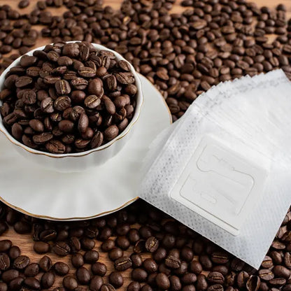 Single serve coffee filters pictured on a bed of coffee beans next to a cup and saucer.  The cup is full of coffee beans.