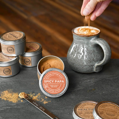 Spicy Papa flavor coffee dust being added by hand to a large mug of hot chocolate. A full-size tin of Healthful coffee dust and several other flavors are depicted on a slate table top.