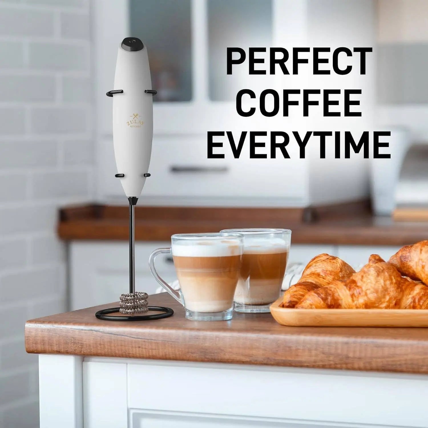 Zulay Tornado Milk Frother depicted upright in its mount on a kitchen counter with two cups of coffee and several croissants.