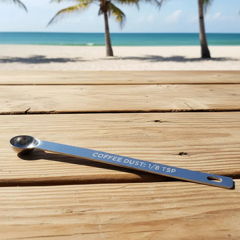 Metal measuring spoon for measuring 1/8 teaspoon of "COFFEE DUST" on a picnic table at a warm beach .